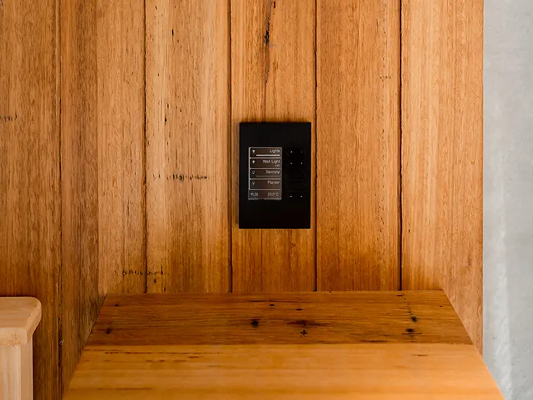 Black wall-mounted C-Bus panel with labeled buttons on a wooden wall.