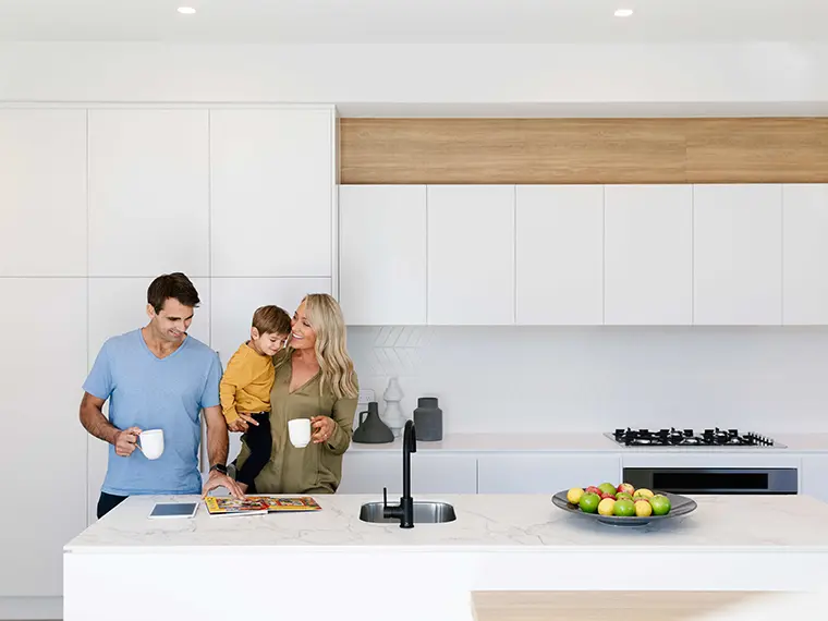 Family in modern white kitchen with island, holding mugs and a baby.