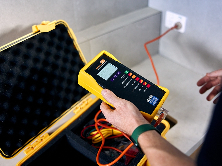 Hand holding a yellow electrical tester over an open case with cables.