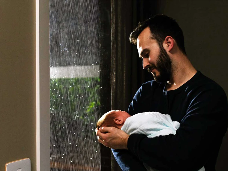 Adult holding a newborn wrapped in a blanket by a rain-covered window with soft natural light.