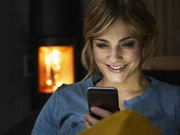 A woman smiles at her smartphone while sitting in a living room.