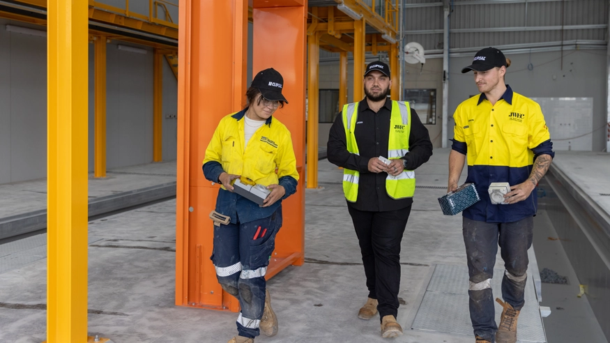 Three men wearing yellow vests and hard hats inside an industrial building, discussing a project while walking.