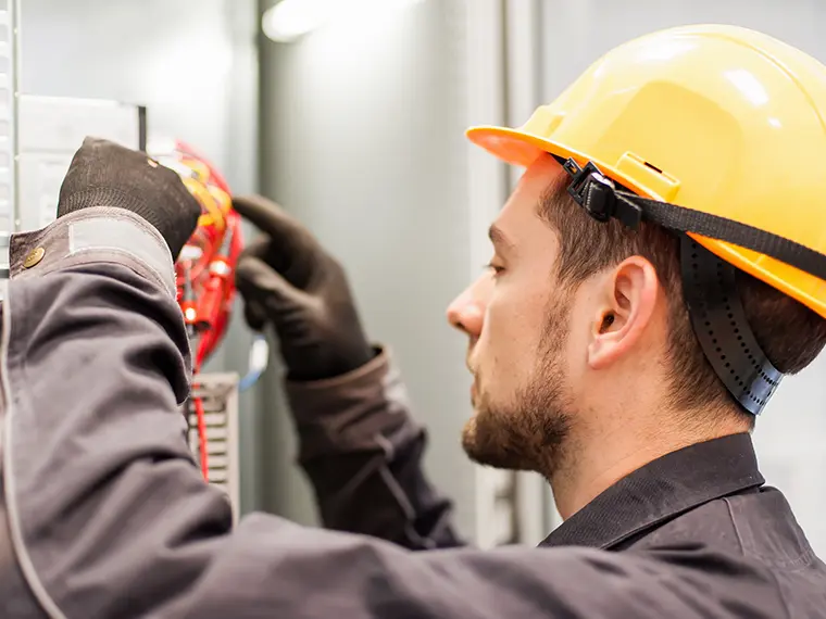 Electrician in yellow hard hat and gloves inspecting components inside an electrical panel.