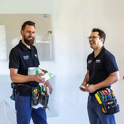 Two tradespeople with tool belts holding electrical supplies in a partially finished room.