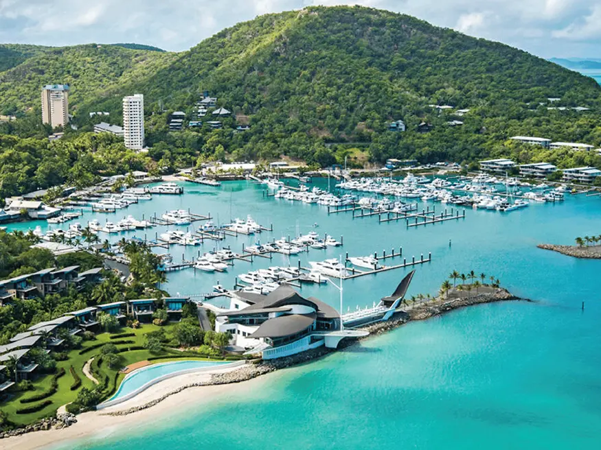 Aerial view of a marina with yachts docked, surrounded by green hills and turquoise water.