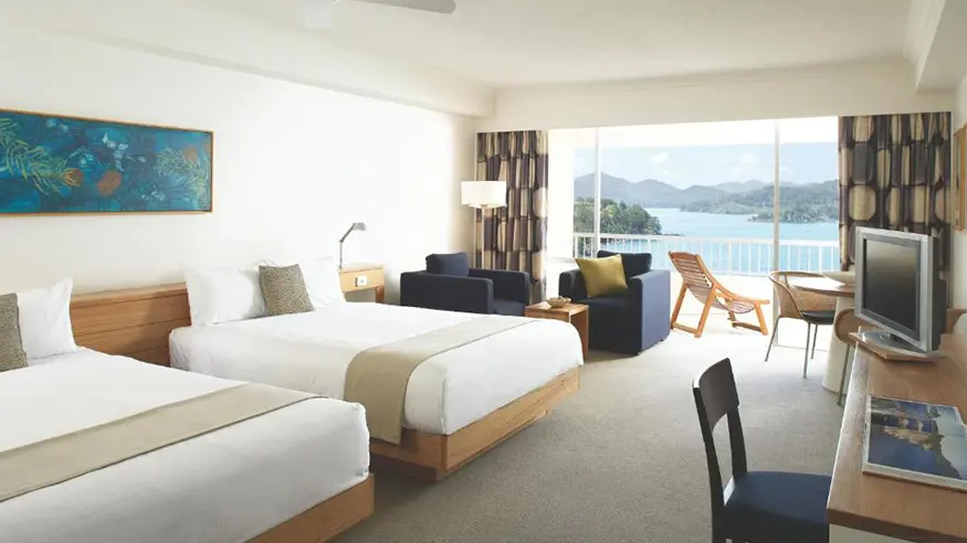 Hotel room with two beds, seating area, desk, TV, and balcony overlooking the ocean and mountains.