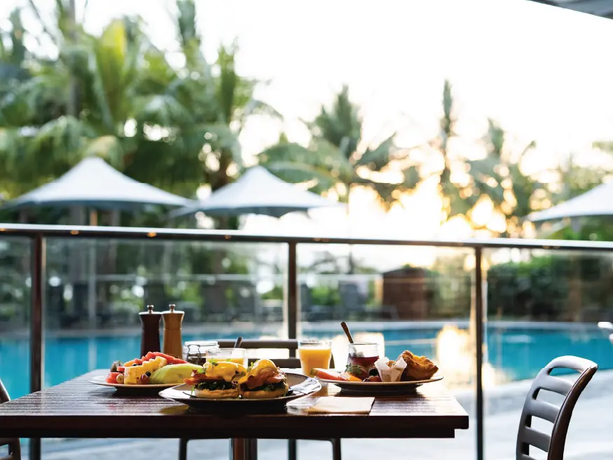Outdoor breakfast table with plates of food, drinks, and a pool in the background at sunrise.