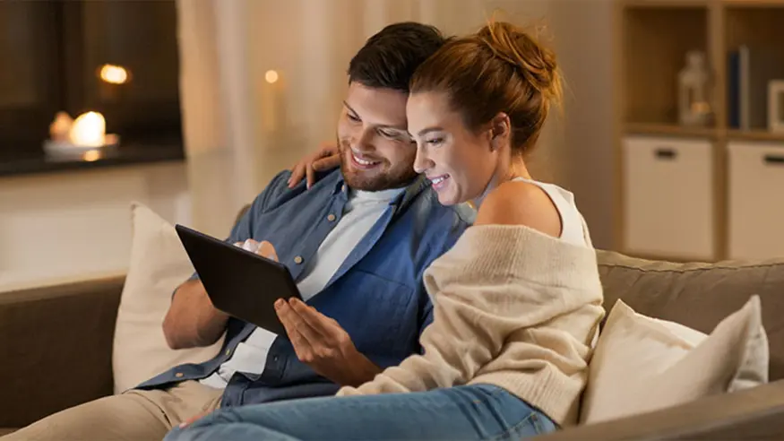 Two people sitting on a couch indoors, one holding a tablet.