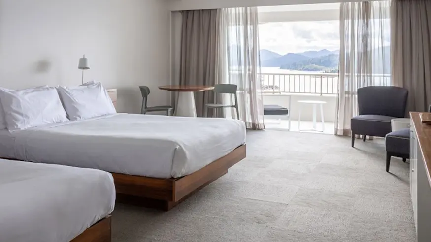Hotel room with two beds, table and chairs, armchair, and window view of ocean and mountains.