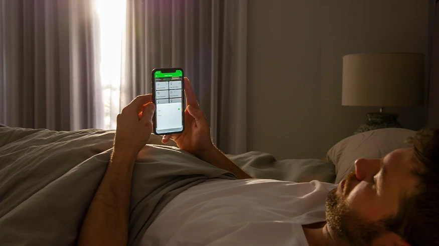 A man relaxing in bed, using his mobile phone to control smart home features via the Clipsal Wiser app.