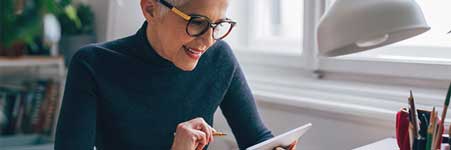 A woman sitting at a desk, wearing glasses and working on a tablet.