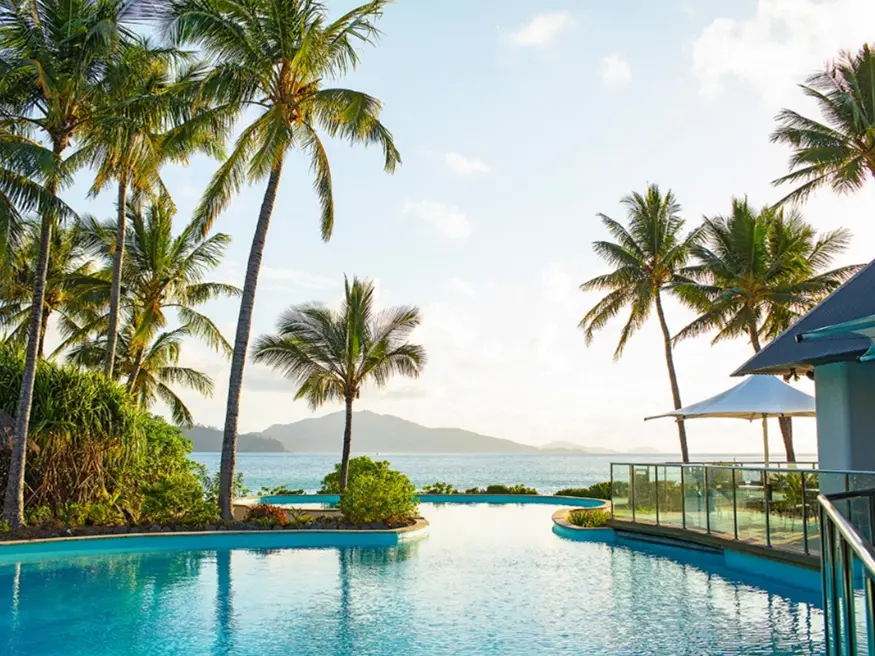 Tropical poolside with palm trees, ocean horizon, and distant mountains under clear sky.