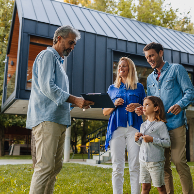 A family of three stands in front of a house as a man shows them a clipboard discussing home design details.