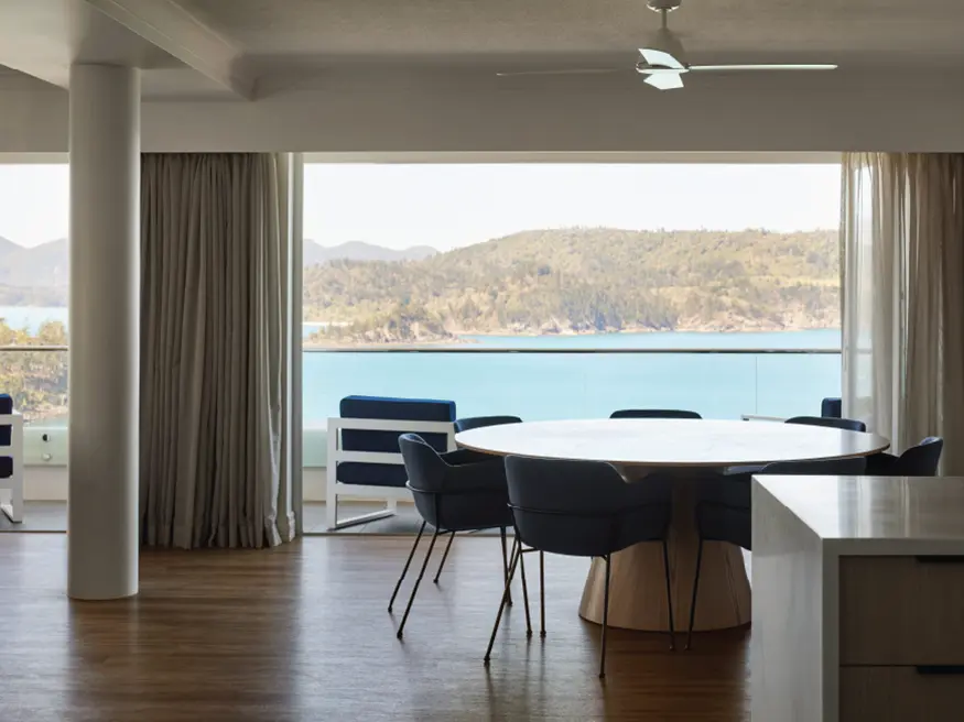 Modern dining room with round table, blue chairs, and large window overlooking a scenic lake and hills.