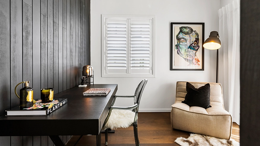 Modern home office with black desk, artwork, cozy lounge chair, and window shutters.