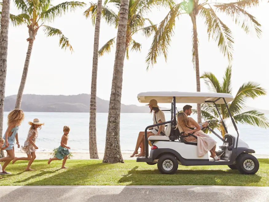 Family with children running near a golf cart on grass by the beach.