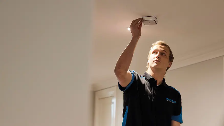 Electrician in dark shirt adjusts a Clipsal Fire Tek smoke alarm on the ceiling.