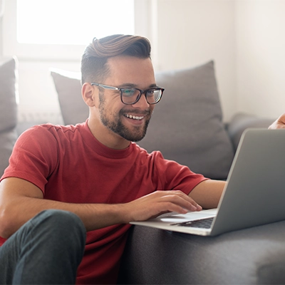 Person in red shirt using a laptop while seated on the floor beside a couch in a casual setting.