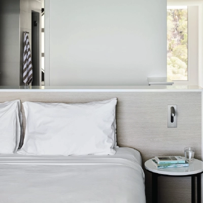 Modern bedroom with white bedding, side table with books and glass of water, and a view into the bathroom.