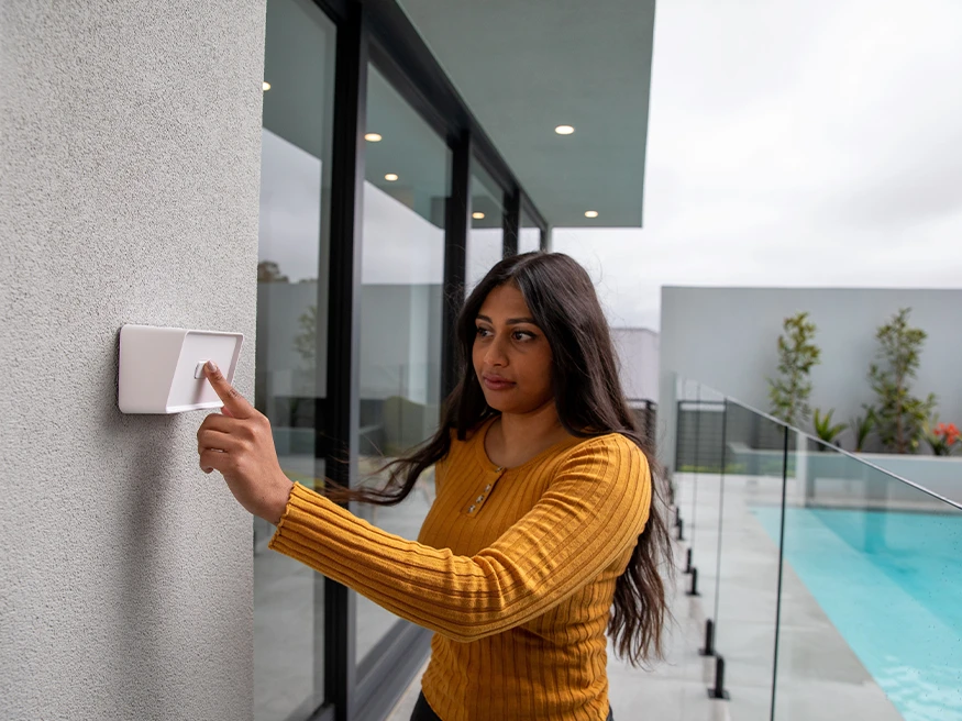 A woman stands near a pool, pointing at a smart thermostat mounted on the wall.
