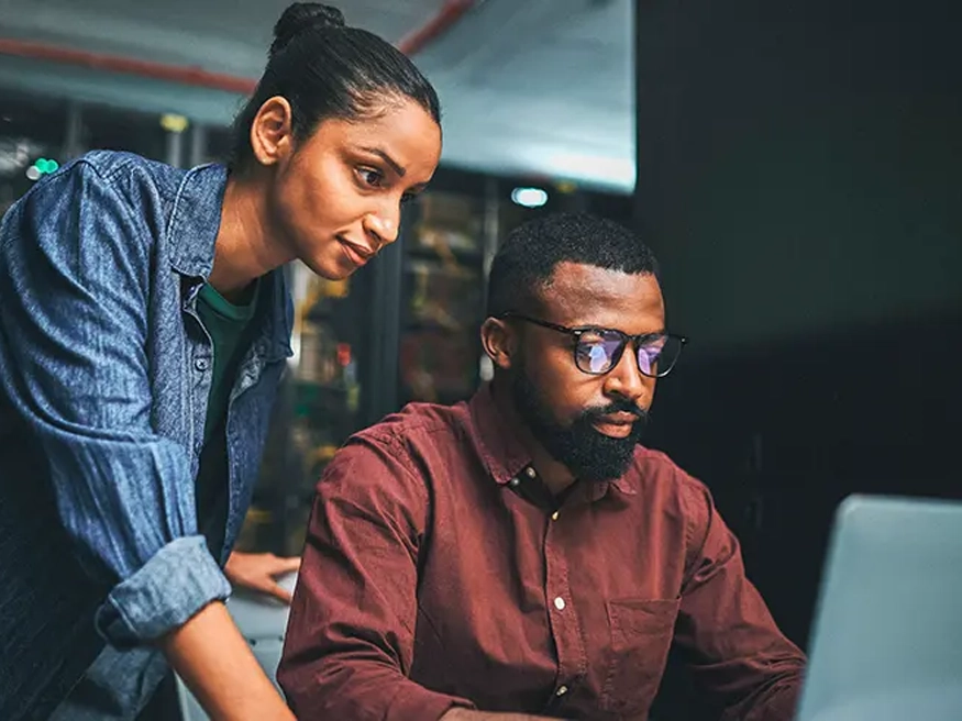 Two people collaborating at a desk, with one seated and typing on a laptop while the other stands and points at the screen.