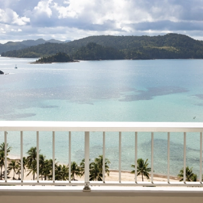 Scenic view of a tropical beach with palm trees, clear blue water, and distant green hills under a partly cloudy sky.