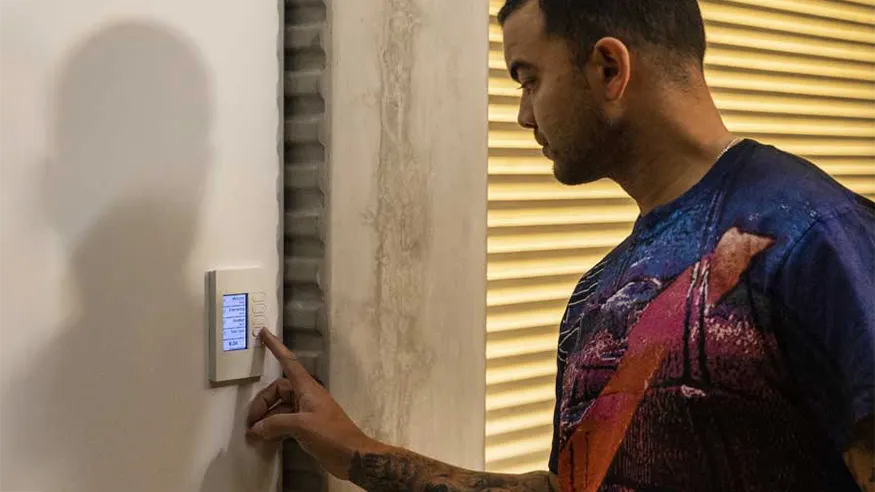Guy Sebastian interacting with a wall-mounted C-Bus control panel, next to a window with closed blinds.