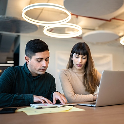 Two people collaborating at a table with a laptop and papers in a modern office setting.