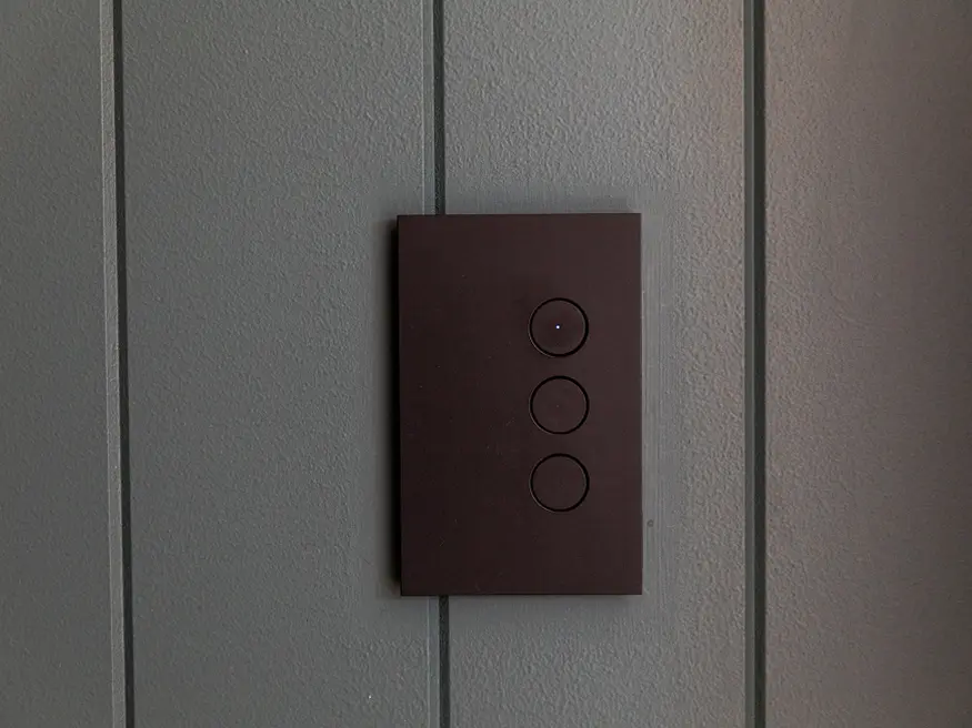 A 3-gang Saturn Zen push button switch in Zen Black colorway mounted on a vertically textured wall.