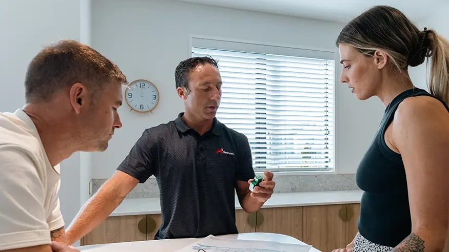 Three people discuss blueprints on a kitchen counter.