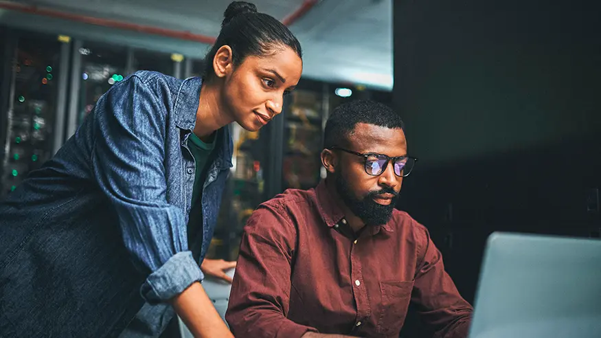 Two people collaborating at a desk, with one seated and typing on a laptop while the other stands and points at the screen.