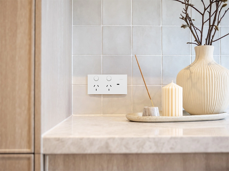 Modern kitchen corner featuring a white power outlet on tiled wall, with a decorative vase, candle, and minimalist tray