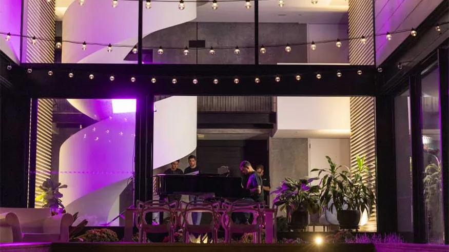 Guy Sebastian’s modern backyard with string lights, potted plants and stylish seating. The indoor space is visible through large glass windows, showing a large white spiral staircase and people gathered around a black grand piano.