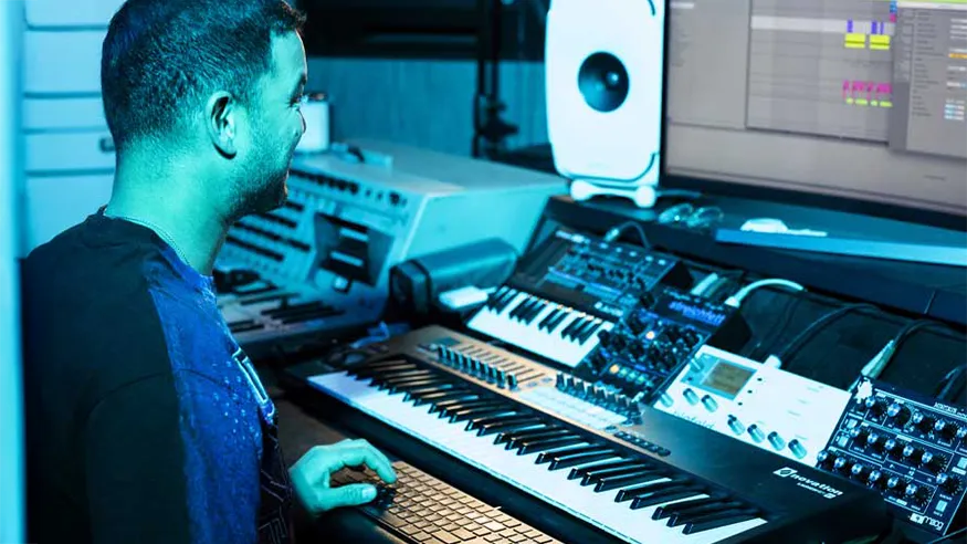 Guy Sebastian working at his in-home music production workstation with a large monitor displaying music software, surrounded by keyboards, synthesisers, and audio equipment.