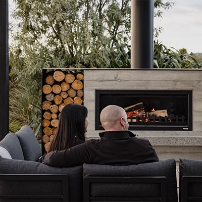 Two people sitting on an outdoor sofa facing a modern fireplace, with stacked firewood and greenery in the background.