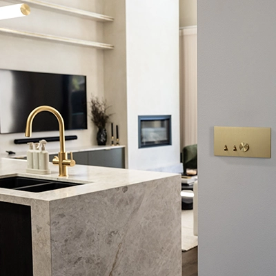 Clipsal Solis brushed brass light switch on wall in modern kitchen with marble island and gold fixtures.