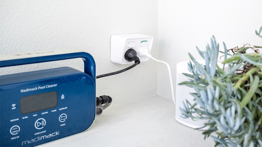 Outdoor setup featuring an Iconic outdoor power point with two plugged-in devices, including a pool cleaner, beside a leafy potted plant.