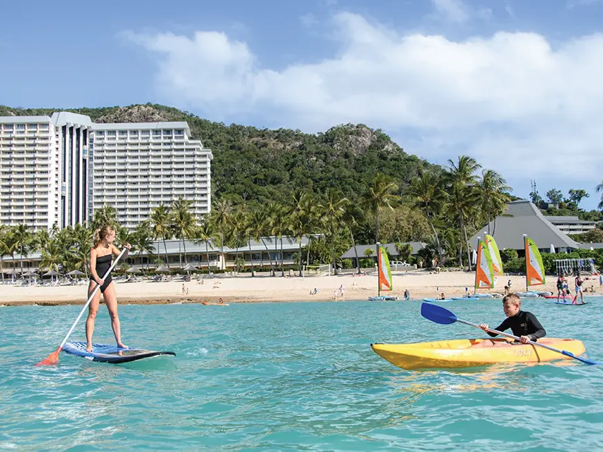 Woman paddleboarding and man kayaking in turquoise water near a beach with hotel, palm trees, and hills behind.