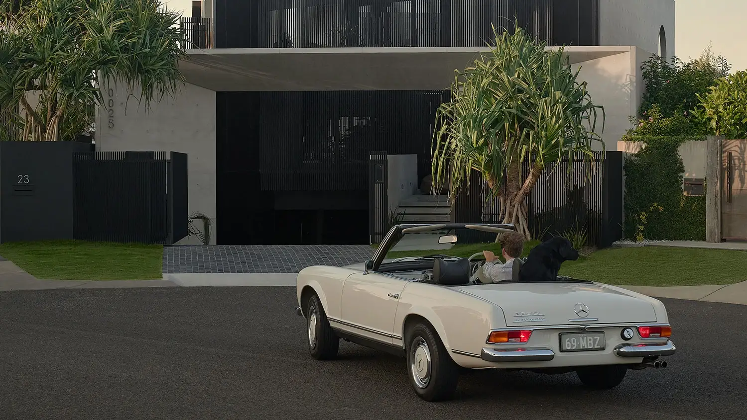 A man and a dog in a white convertible car parked in front of a modern house with black gates.
