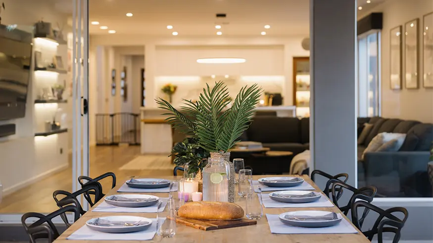 Dining table set for six with bread, candles, and palm leaves centerpiece in a modern open-plan living space.