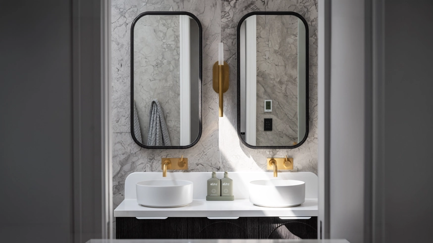 Modern bathroom with marble walls, black-framed mirrors, gold faucets, and a central light fixture, highlighting a stylish home renovation. A black Iconic Styl can be seen through one of the mirrors.